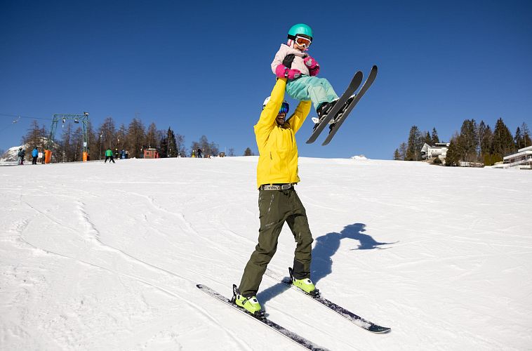 Vater trägt Kind auf den Schultern beim Skifahren. Blauer Himmel und verschneite Piste.