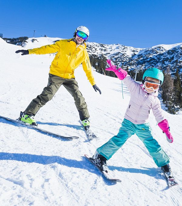Erwachsener und Kind beim Skifahren auf einer sonnigen, schneebedeckten Piste.