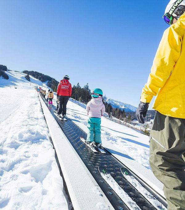 Kinder und Erwachsene auf einer Skipiste im Schnee, bei klarem Himmel, während sie Ski fahren.