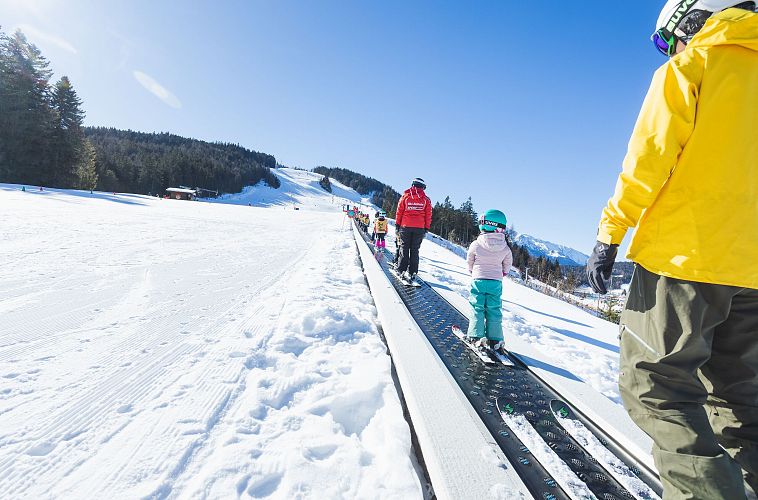 Kinder und Erwachsene auf einer Skipiste im Schnee, bei klarem Himmel, während sie Ski fahren.