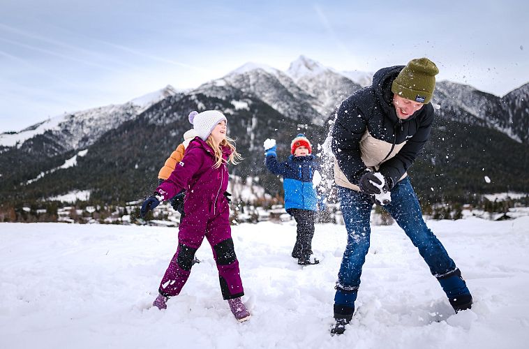 Drei Personen im Schnee vor Bergen in der Region Seefeld. Kinder werfen Schnee in die Luft. Winterkleidung, blauer Himmel.