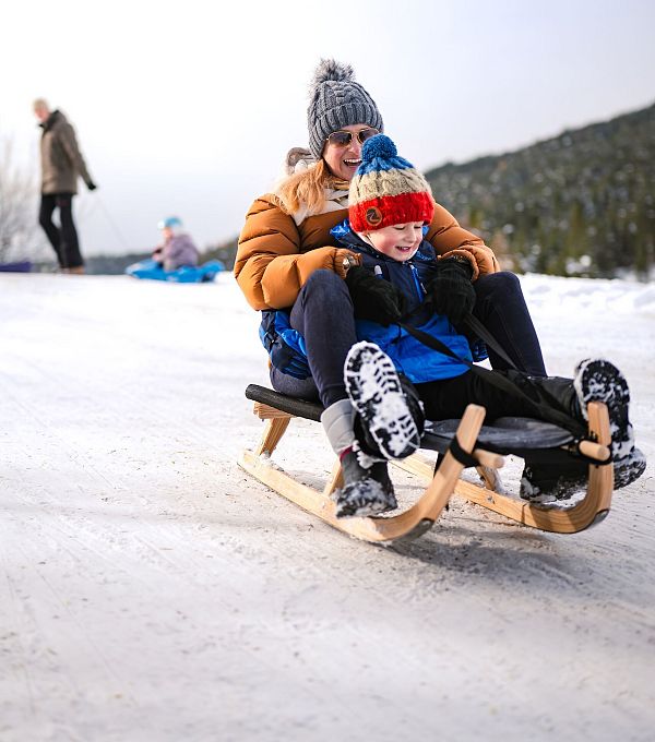Una madre e un bambino su una slitta sulla neve, sorridendo e indossando abbigliamento invernale.