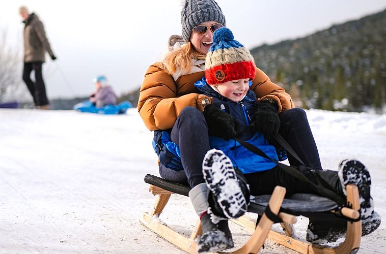 Frau und Kind rodeln auf einem verschneiten Hügel. Winteraktivitäten in der Region Seefeld.