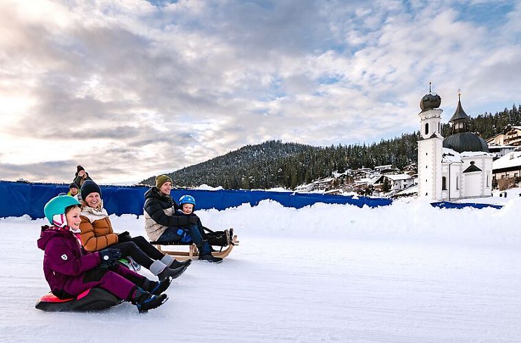 Menschen rutschen auf einer Eisfläche, im Hintergrund eine verschneite Kirche. Winteraktivität in der Region Seefeld.