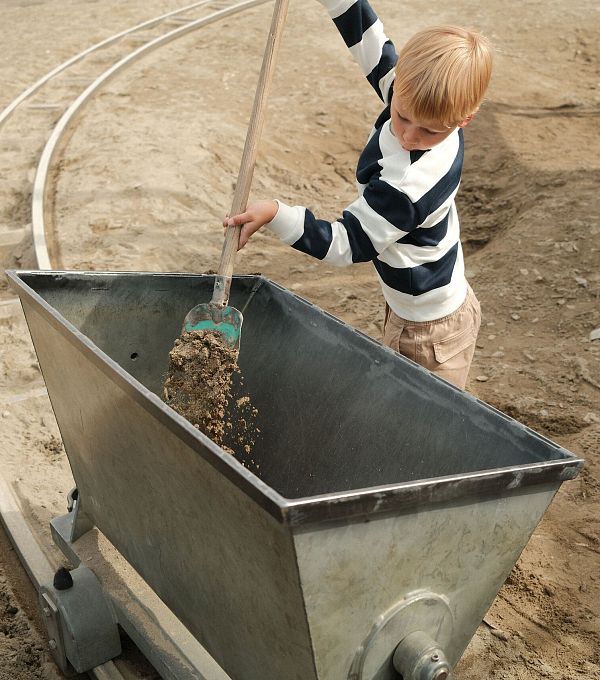A boy with a striped shirt shovels gravel into a metal container on a playground.