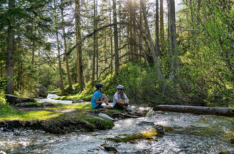 Zwei Radfahrer sitzen an einem Bach im Wald, umgeben von Bäumen. Die Sonne scheint durch das Blätterdach.