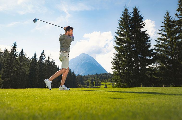 Ein Mann spielt Golf auf einem grünen Platz mit Bergen im Hintergrund.