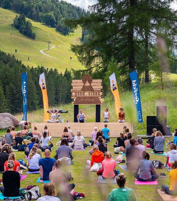 Menschen sitzen auf der Wiese und beobachten eine Aufführung auf einer Bühne in der Natur.