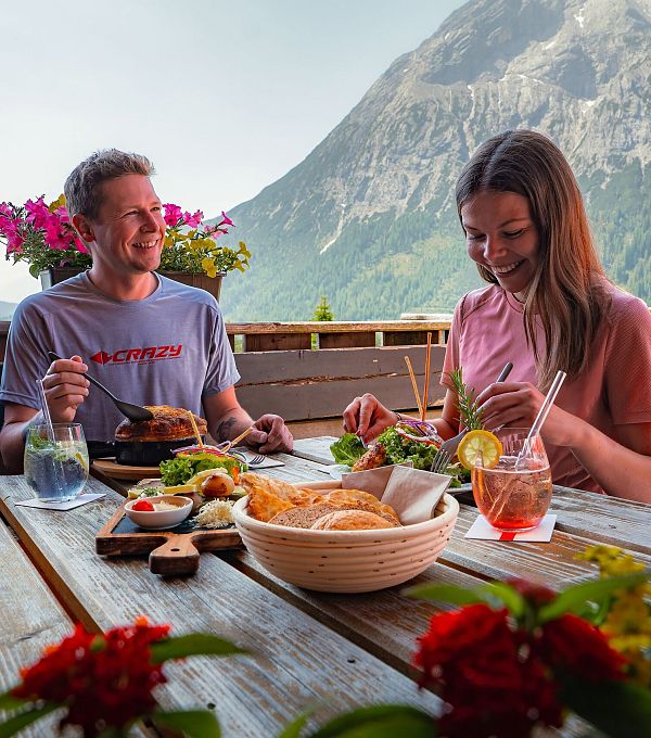 Paar beim Essen im Freien, mit Bergen im Hintergrund und blühenden Pflanzen am Tisch.