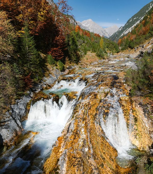 Bergfluss mit Kaskaden in herbstlicher Kulisse und schneebedecktem Gipfel im Hintergrund.