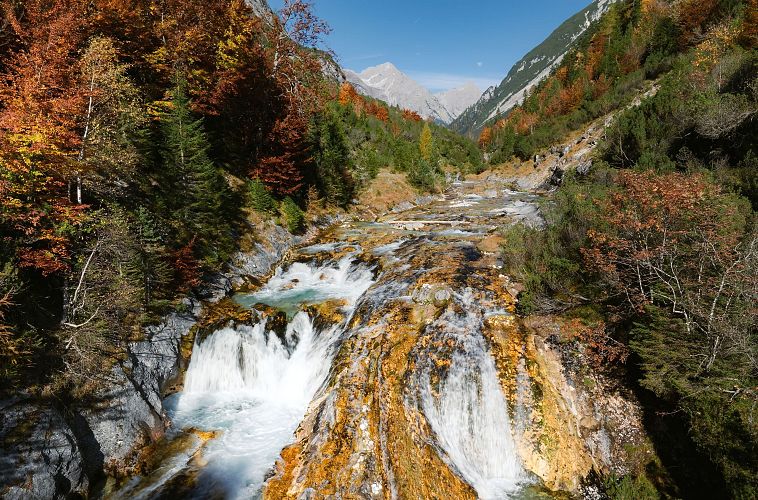 Bergfluss mit Kaskaden in herbstlicher Kulisse und schneebedecktem Gipfel im Hintergrund.