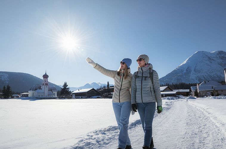 Zwei Frauen wandern im Schnee in der Region Seefeld, mit Kirche und Bergen im Hintergrund.