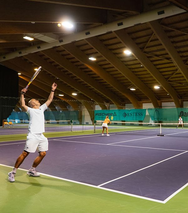 Tennishalle, Spieler in weiß serviert. Weitere Spieler im Hintergrund aktiv. Helle Beleuchtung. Innenaufnahme.