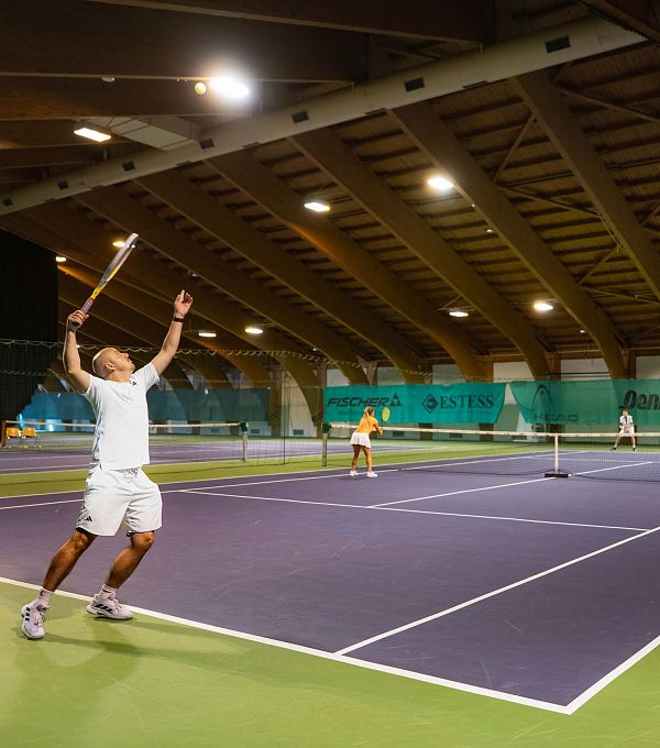 Mehrere Personen spielen Tennis in einer Indoor-Anlage, ein Spieler serviert.