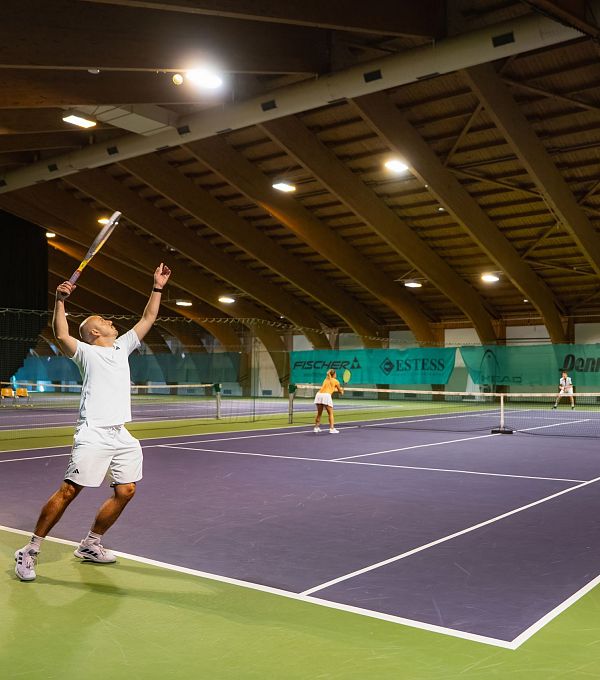 Mehrere Personen spielen Tennis in einer Indoor-Anlage, ein Spieler serviert.