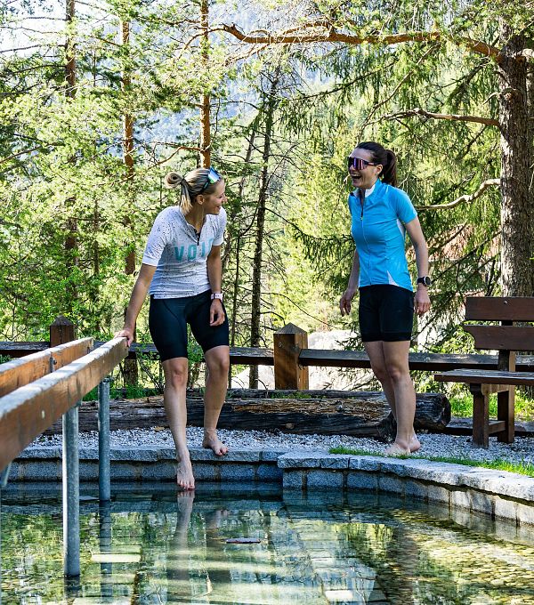 Zwei Frauen stehen an einem Teich in der Natur, eine mit Füßen im Wasser, eine auf dem Geländer.