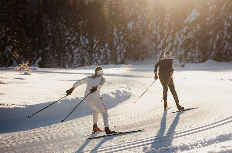 Zwei Personen beim Langlaufen auf einer schneebedeckten Strecke in der Sonne.