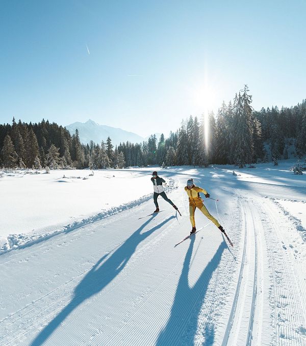 Zwei Langläufer auf einer verschneiten Spur in der Region Seefeld, umgeben von Bäumen und strahlendem Sonnenschein.