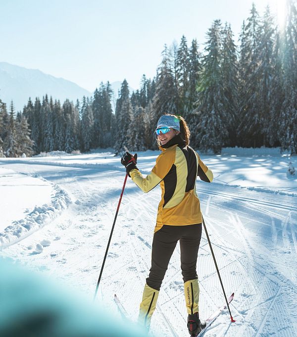 Langläuferin in gelb auf sonniger Loipe in der Region Seefeld, umgeben von verschneiten Bäumen.