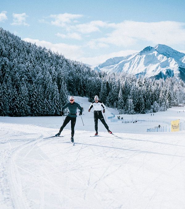 Zwei Langläufer auf einer schneebedeckten Langlaufpiste mit Bergen im Hintergrund.