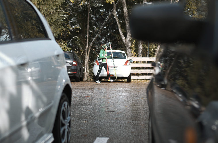 Frau mit Wanderstock auf Parkplatz, umgeben von Autos. Bäume und Holzzaun im Hintergrund. Saison Herbst.