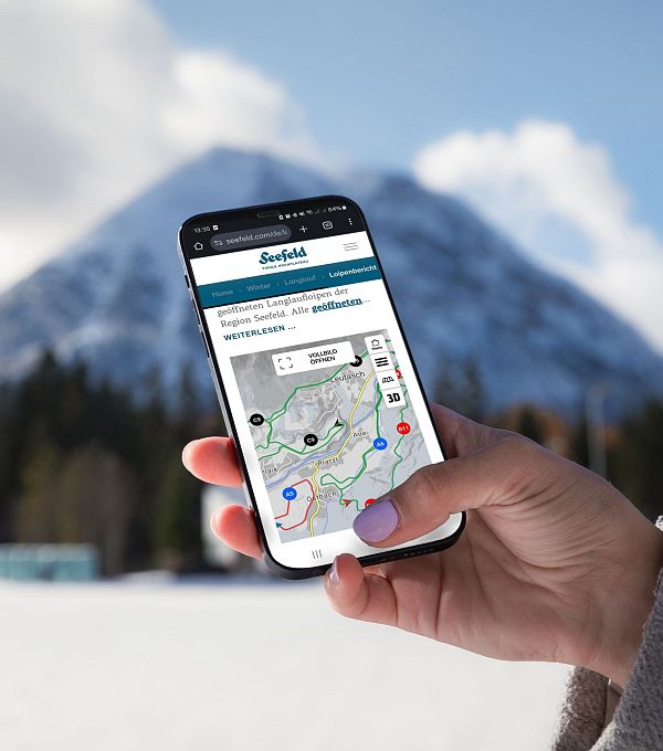 Hand hält Smartphone mit Karte der Region Seefeld. Dahinter schneebedeckte Berge und blauer Himmel.