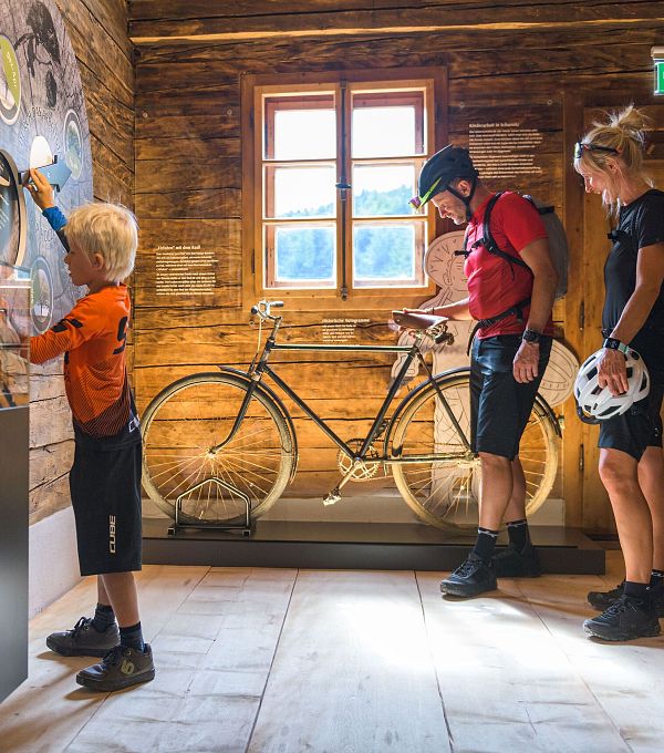 Ein Junge interagiert mit einer Wandtafel, während zwei Erwachsene ein Fahrrad betrachten.
