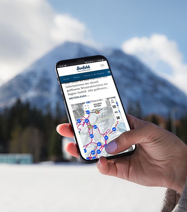 A hand holding a smartphone with a winter hiking map, snow-covered mountains in the background.