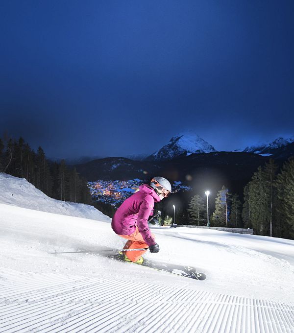 Two skiers on a snowy slope at night, with bright lights and mountains in the background.