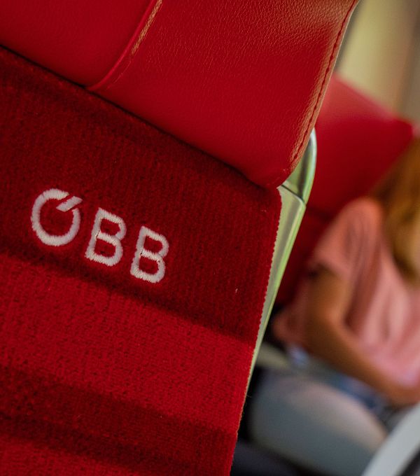 A red train seat with the ÖBB logo and a woman sitting in the background.