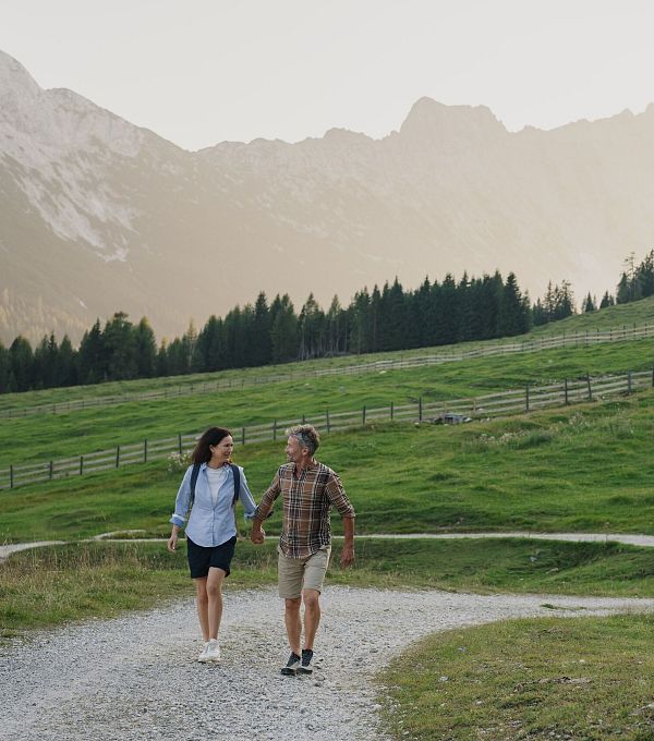 Paar geht Hand in Hand auf einem Weg in einer grünen Landschaft mit Bergen im Hintergrund.