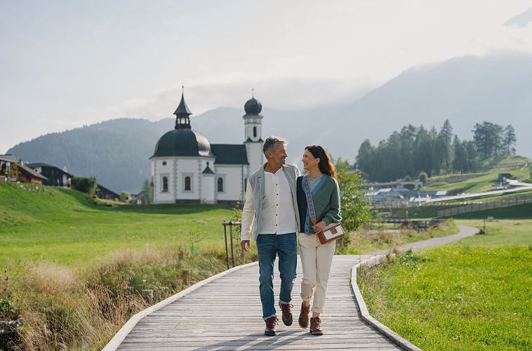 Paar spaziert vor Kirche in der Region Seefeld auf hölzernem Steg. Im Hintergrund Berge und Wiesen.