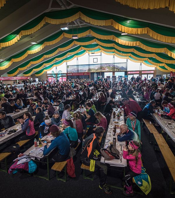 Großer Veranstaltungsraum mit Menschen an langen Tischen unter einem Dach mit gelb-grünen Stoffbahnen, lebendige Atmosphäre, viele Besucher.