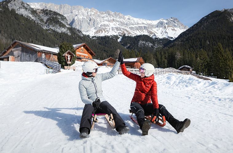 Zwei Personen auf Schlitten im Schnee vor Bergen und Hütten in der Region Seefeld.