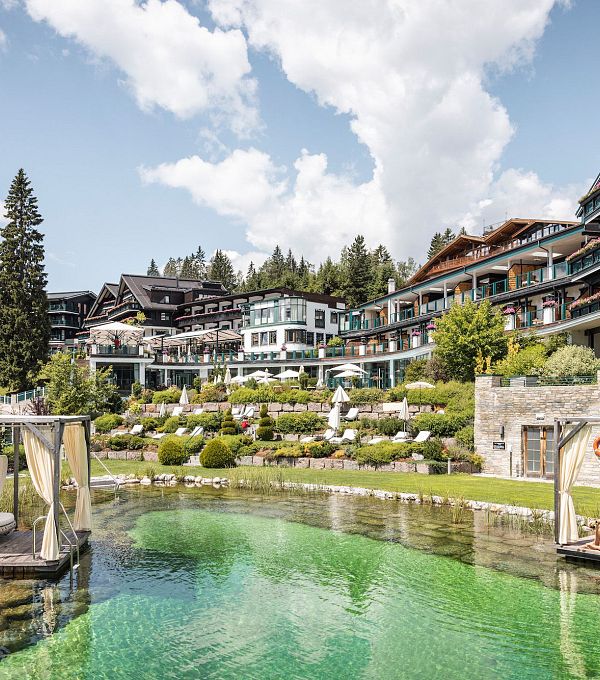 View of a modern resort by a green pond surrounded by trees and blue sky.