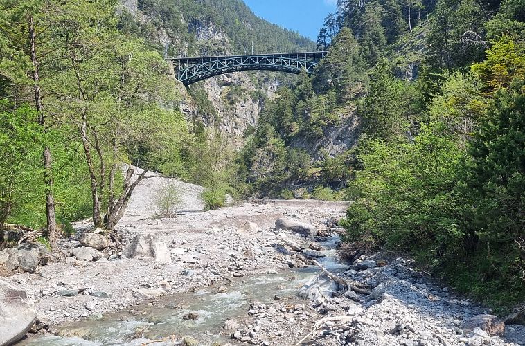 Ein Fluss fließt unter einer Eisenbrücke, umgeben von Felsen und Bäumen.