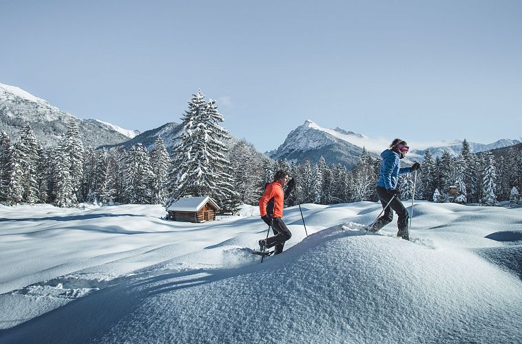 Zwei Personen beim Schneeschuhwandern in der verschneiten Region Seefeld, mit Bergen und Hütte im Hintergrund.