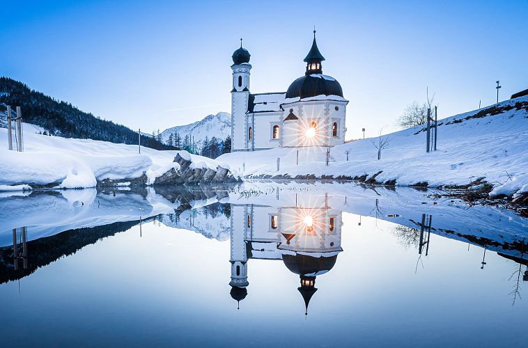 Winter landscape with a church reflected in still water, snow-covered ground and clear blue sky.
