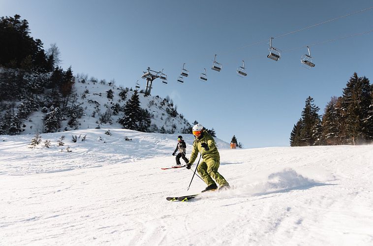 Ein Skifahrer in grüner Kleidung fährt die Skipiste hinunter, im Hintergrund Skilifte.