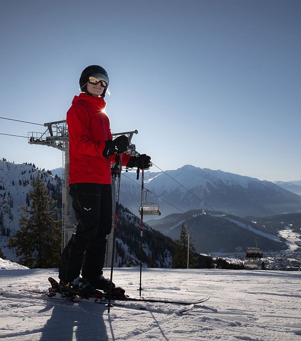 Skier in a red jacket on snowy slopes with mountains and a ski lift in the background.
