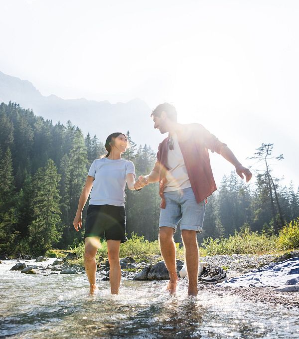 Couple walking barefoot in a river, surrounded by trees and mountains with sunlight shining.