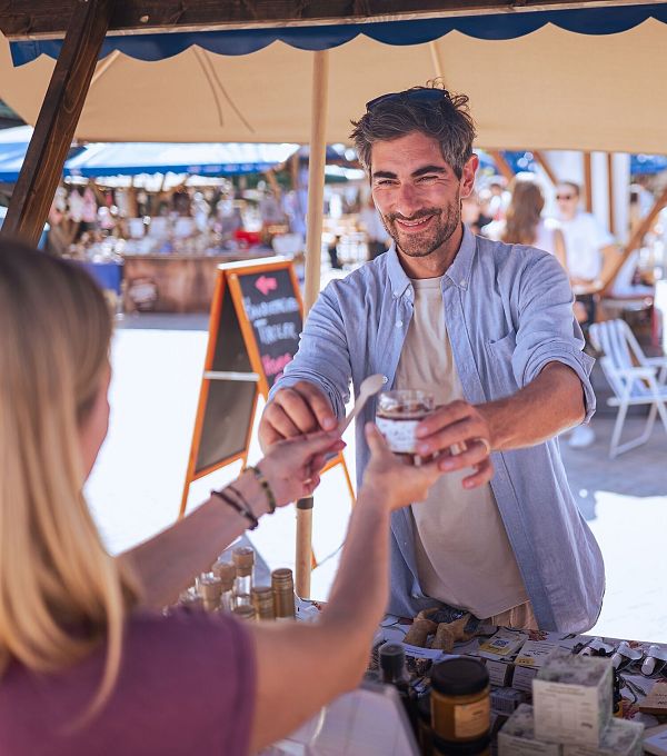 A customer pays a vendor at an outdoor market. A woman stands beside them looking happy.
