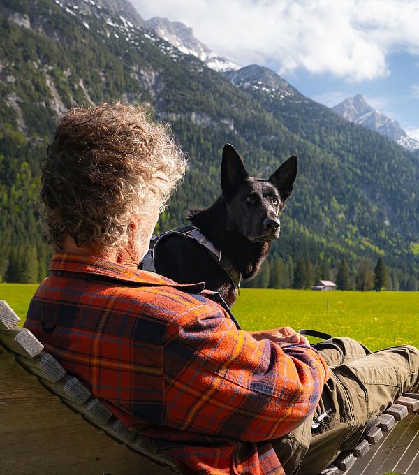 Ein Mann sitzt mit einem Hund auf einer Liege vor einer Berglandschaft.