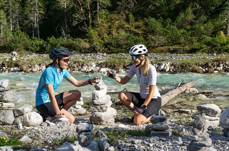 Two women in cycling gear build stone towers by a river.