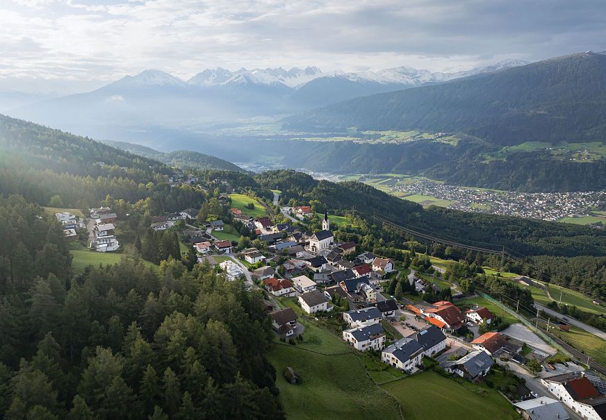 Luftaufnahme eines kleinen Dorfes in bergiger Landschaft mit Hügeln und Bergen im Hintergrund.