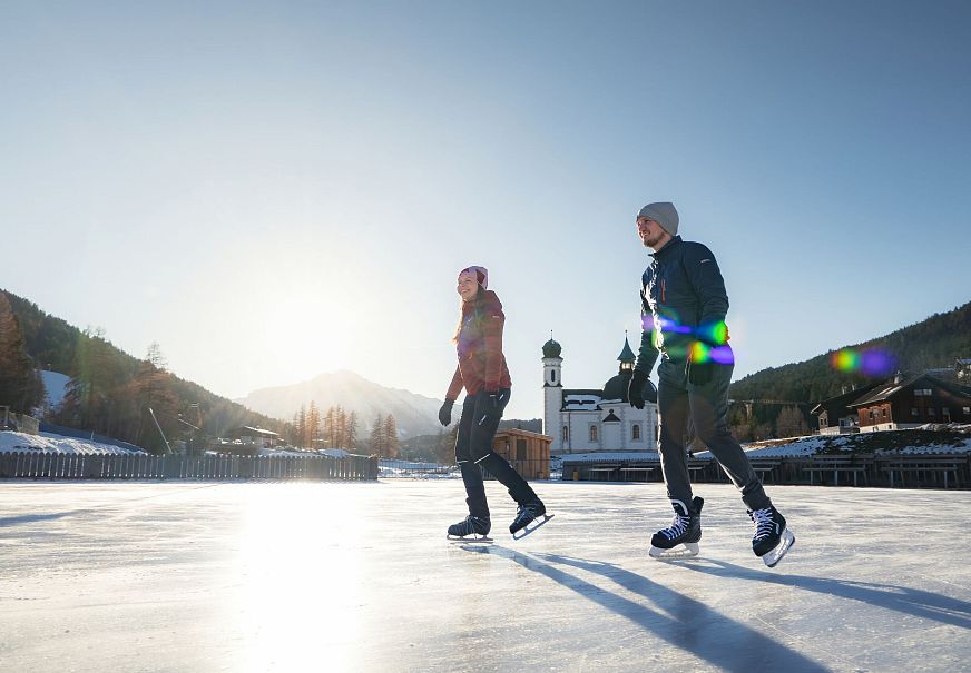 Zwei Menschen eislaufen draußen unter blauem Himmel. Kirche im Hintergrund. Wintersport in der Region Seefeld.