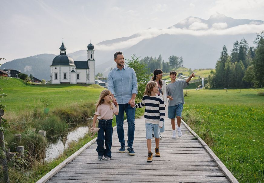 Eine Familie geht auf einem Holzsteg in einer grünen Landschaft mit einer Kirche im Hintergrund.