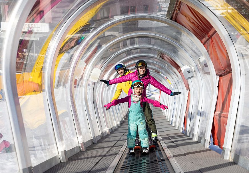 Drei Kinder stehen fröhlich auf einem Förderband im Schnee, umgeben von einer transparenten Struktur.
