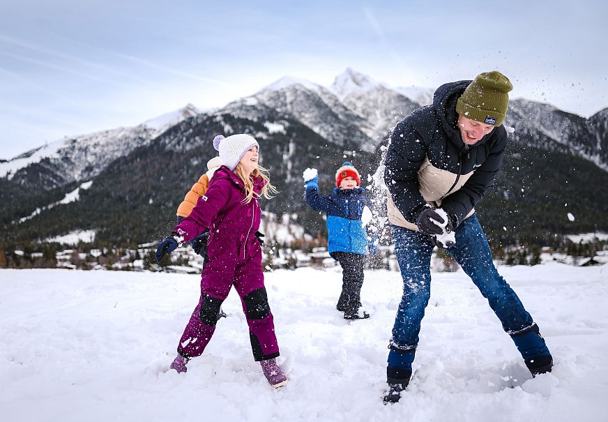 Drei Personen im Schnee vor Bergen in der Region Seefeld. Kinder werfen Schnee in die Luft. Winterkleidung, blauer Himmel.