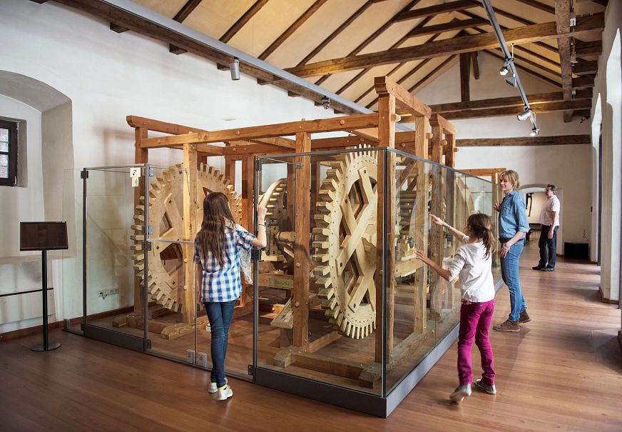 Visitors looking at a wooden gear mechanism in a museum exhibit.